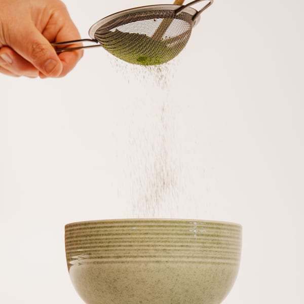 A hand sifting green matcha powder into a light green ceramic bowl.