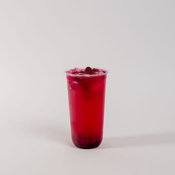 A vibrant red drink with ice and fruit pieces in a clear plastic cup on a white background.