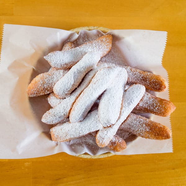 Basket of golden-brown fried pastries heavily dusted with white powdered sugar on a wooden table.