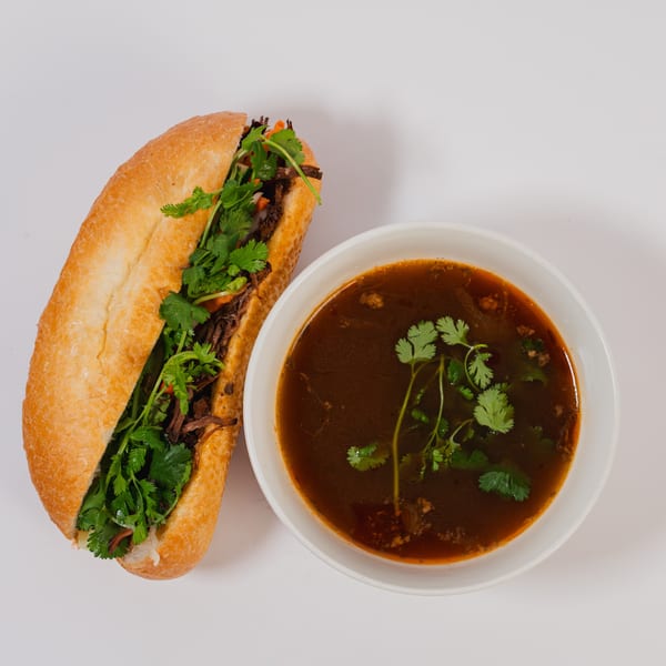 Banh mi sandwich filled with meat and cilantro, served alongside a bowl of dark broth soup.