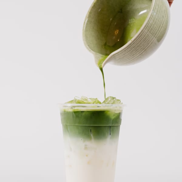 Matcha green tea being poured from a bowl into an iced latte on a white background.