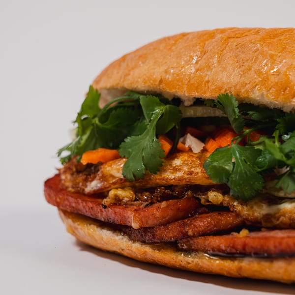 Close-up of a Banh Mi sandwich with fried eggs, sliced meat, cilantro, and pickled carrots.