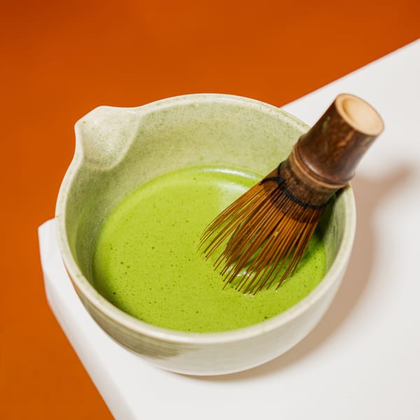 A light green bowl of whisked matcha tea with a bamboo whisk, on a white surface with an orange background.